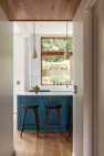 A peek from the hallway into a kitchen with wood stools at the counter and a large window beyond.