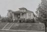 A historic black and white photograph of a single-story home. It's viewed from the street and situated slightly up on a berm.