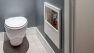 A wall-mount toilet in a grey-blue bathroom with a grey tile floor. There's a magazine holder built into the wall.