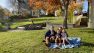 Ian sits on a picnic blanket with his wife and three children. They're on a lawn in front of some historic buildings. The sun is out and they're smiling.