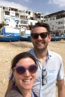 Lindsey and her husband stand on the beach taking a selfie. There are docked boats and building in the background, all in white and blue.