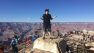 Malcom stands on a large rock overlooking the Grand Canyon. It's a clear, sunny day. He's gesturing away from the camera with his arms out at his sides.