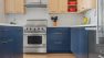 Looking into a bright, clean kitchen. The lower cabinets are slate blue and the uppers are light wood. The walls are clad in white subway tile and there are orange pots on the shelves.