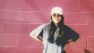 Sneha stands in front of a red brick wall. She's wearing a white hat and grey t-shirt.