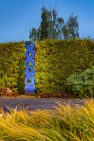 At dusk, a tall yew hedge backdrops a tall sculpture made up of thick silver disks. Some disks have organically shaped holes cut out of them, like donuts. The sculpture and hedge are illuminated from below.