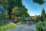 Looking across the bluestone patio toward the back of the garden. Steps up the deck and to the left and a decorative sculpture peeks out at the right. In the center sits a light-wood table and chairs.