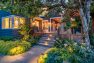 Dusk outside a blue home with a large raised port illuminated by exterior and interior lights. The garden surrounding the porch is verdant and green. — Residential Landscape Architecture by Board & Vellum
