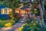 Dusk outside a blue home with a large raised port illuminated by exterior and interior lights. The garden surrounding the porch is verdant and green. — Residential Landscape Architecture by Board & Vellum