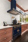 A white kitchen with light wood cabinets. There is a large range and hood at the center, which is navy blue with gold knobs and pulls.