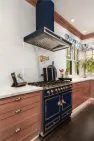 A white kitchen with light wood cabinets. There is a large range and hood at the center, which is navy blue with gold knobs and pulls.