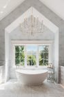 An all grey-and-white bathroom with a large soaking tub. There is a window behind the tub looking out at trees. It is a sunny day and there are sun rays illuminating the bathroom.