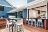 A hightop table with hightop chairs is at the center of a wood deck. Behind is a built-in grill with a sink and countertop. To the right is a large bar top with chairs that looks into the kitchen via a large, operable window.