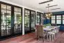 An outdoor dining table seating eight is arranged on a wooden deck. The area is covered by a roof where lighting and radiant heating are inset. Behind are a series of French doors leading to the indoor dining room.