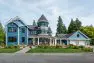 A large blue house with a gently sloping lawn and prim plantings around the perimeter. The house is large with a large turret and wrap around deck.