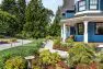 A cottage-style garden with an array of plants in many colors and textures. Behind is a bright blue house, walking path, and lawn.