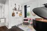 A collection of electric and acoustic guitars and ukuleles hang on a white wall. To the right is the back of a baby grand piano with the lid open.