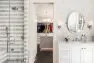 To the left is a grey and white tiled shower. To the right, a single-sink vanity all in white. At the center is a look into the walk-in closet with a center island and crystal chandelier.