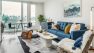 A bright white living room with a feature couch in blue, a coffee table, and a small dining area.