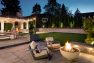 A fire pit with two freestanding stairs stand on a patio. Beyond, a turf lawn connects to a sport court.