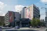 An architectural rendering of the Tanager Apartments looking at the building from street-level at the corner of Madison and 14th. Chop Suey, a well-known music venue, is in front. Behind stands the two towers of the Tanager Apartments. On the south tower façade is an example of a mural intended to enliven the building}s exterior. On the north tower is a sample mural of Freddie Mercury.