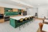 A photo of the interior of a multifamily residential amenity kitchen. Focuses on a large six seat kitchen island with a sink and six black and woven cane barstools. Includes wooden shade pendant lights, wood tone kitchen cabinets, gray tile flooring, and emerald green square tile backsplash.