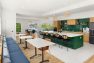 A photo of the interior of a multifamily residential amenity kitchen. Focuses on a large six seat kitchen island with a sink and six black and woven cane barstools. Includes wooden shade pendant lights, wood tone kitchen cabinets, gray tile flooring, and emerald green square tile backsplash. Also includes upholstered banquette seating, bistro tables, and brown leather dining chairs.