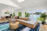 A photo of the interior of a multifamily residential amenity lounge. Focuses on a large upholstered modular sofa seating and large windows with a view of a lake. Includes a large abstract blue rug, a round wooden coffee table, house plants, and a dining set.