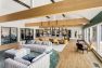A photo of the interior of a multifamily residential amenity clubhouse with vaulted ceilings framed with wide wooden beams. Presents a wide view of the room to show how it has been furnished into several zones with different purposes such as conversation, working, dining, and relaxing.