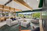 A photo of the interior of a multifamily residential amenity clubhouse with vaulted ceilings framed with wide wooden beams. Focuses on a conversation space furnished with wide, upholstered sofas, a thick rug, and a tiled fireplace.