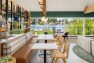 A photo of the interior of a multifamily residential amenity clubhouse with vaulted ceilings framed with wide wooden beams. Focuses on a seating banquette and several bistro tables with wood and dining chairs that are positioned with a view of a lake outside large windows.