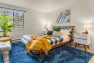 An interior photo of an apartment bedroom. Focuses on a wooden queen sized bed dressed in black check bedding and textured pillows. Includes a large, high pile blue rug, a house plant, abstract art on the walls, and white mid-century nightstands.