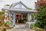 An exterior view of a Victorian style, two level house, focusing on the large, covered porch and the large sliding glass door leading into the house. There are many plants, flowers, and trees in the garden around the house. Judkins Park Victorian – Board & Vellum