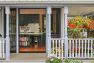 An exterior view of a Victorian style, two level house, focusing on the large, covered porch and the large double French door leading into the kitchen from the porch. There are many plants, flowers, and trees in the garden around the house. Judkins Park Victorian – Board & Vellum