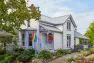 An exterior view of a Victorian style, two level house, focusing on the front entry. The front of the house has a bay window and a rainbow flag. There are many plants, flowers, and trees in the garden around the house. Judkins Park Victorian – Board & Vellum