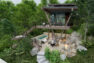 An architectural rendering of the exterior of a treehouse rental property. The rear exterior has a large deck off the home, along with a patio at the lower level, connected via stairs.