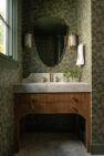 A bathroom vanity in medium wood with a quartz countertop. There is an oval mirror and wall sconces behind the vanity and a floral green wallpaper on the walls.