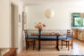 An image of dining room interior, focused on an oval wooden dining table and an upholstered, built-in dining bench. The bench features a blue cushion for the seat and an embroidered floral on white cushion for the backrest. The image also shows a large paper globe pendant light and a ladder back dining chair. On the right, there is a peek of the doorway to the green and white kitchen.