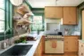 Interior view of a sunlit kitchen showcasing maple and apple plywood cabinets, white marble countertops, and walls with green trim and an arts and crafts style green wallpaper.