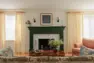 A wide interior view of a living room, with a fireplace in the center and a large window on either side. The windows are dressed with cream fabric drapes and white sheer roman shades. Also shows parts of furnishings including a pink velvet chair, an antique wood coffee table, and a floral patterned sofa.