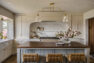 A traditional-style kitchen in a single-family home with ivory-painted cabinetry, a white and gray quartz countertop, and brass hardware. There is a large central, dark wood island sits in front of the gas range with three upholstered seats.