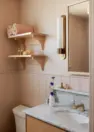 An interior view of a small bathroom, focused on the light pink walls. Also features wood open shelving, a vertical, cylindrical brass and white glass wall sconce, a wood framed mirror, and a sink with a marble countertop and brass hardware.
