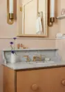 An interior view of a small pink bathroom, focused on the area around the sink. The sink features a marble countertop and backsplash with an elevated lip, brass faucet and hardware, and a maple and apple plywood vanity cabinet below. Also shown in the image is a light wood framed rectangular mirror and two brass and white glass cylindrical wall sconces.
