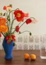 An image of dining room interior, focused on the surface of a wooden dining table and an embroidered floral on white backrest of a dining bench. The image also shows a floral arrangement of red, orange, pink, and white poppies.