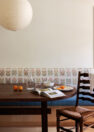 An image of dining room interior, focused on an oval wooden dining table and an upholstered, built-in dining bench. The bench features a blue cushion for the seat and an embroidered floral on white cushion for the backrest. The image also shows a large paper globe pendant light and a ladder back dining chair.