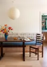 An image of dining room interior, focused on an oval wooden dining table and an upholstered, built-in dining bench. The bench features a blue cushion for the seat and an embroidered floral on white cushion for the backrest. The image also shows a floral arrangement of colorful poppies, a large paper globe pendant light and a ladder back dining chair. On the right, there is a peek of the doorway to the green and white kitchen.