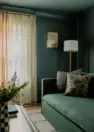 An interior view of a a small multipurpose room that focuses on a simple dark green sofa and green walls and ceiling. Includes a window with light filtering cream drapes with rickrack trim.