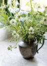 A vignette image of a green and white floral arrangement in an antique metal pitcher situated by a window.