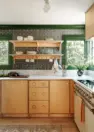 Interior view of a sunlit kitchen showcasing maple and apple plywood cabinets, white marble countertops, and walls with green trim and an arts and crafts style green wallpaper.