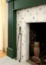 An image focused on part of a fireplace with a green wooden mantle, a white marble hearth, and a white and blue Delft tile surround. Includes iron tools including a fireplace grate with a an owl sculpture.