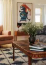 An interior view of a living room, with focus on a wooden coffee table. Also includes a blue and white shag rug, a wooden console table, a red metal lamp, and an abstract painting of a bright orange and turquoise figure.